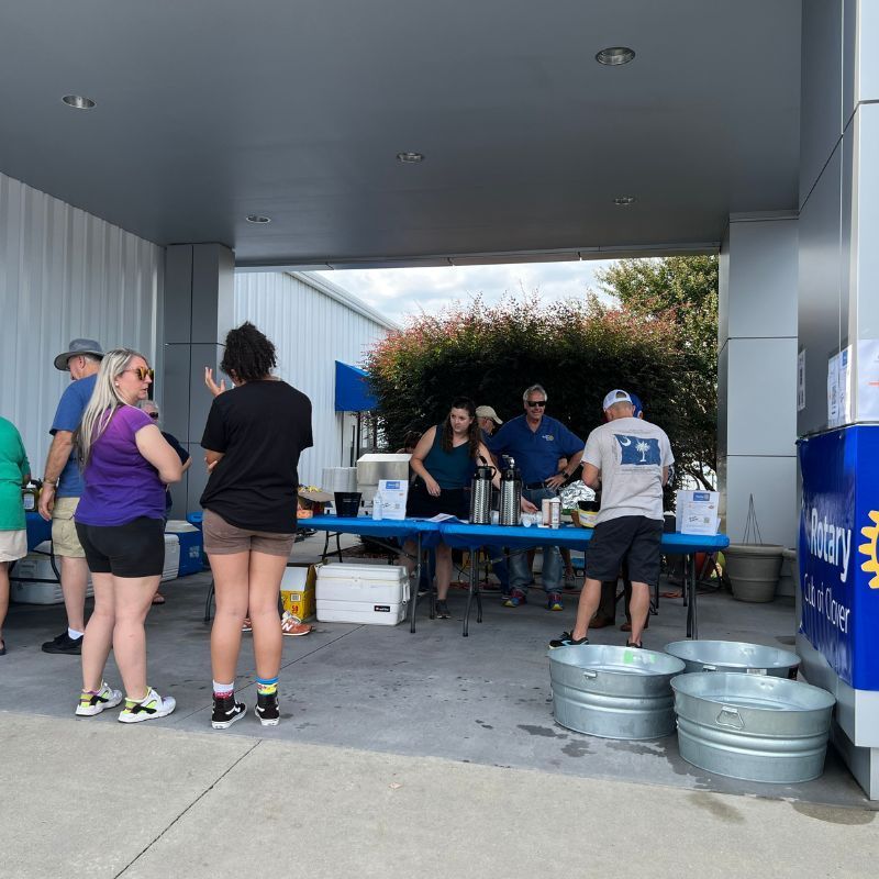 People serving drinks at a table under a covered outdoor area, with coolers and beverage kegs nearby.