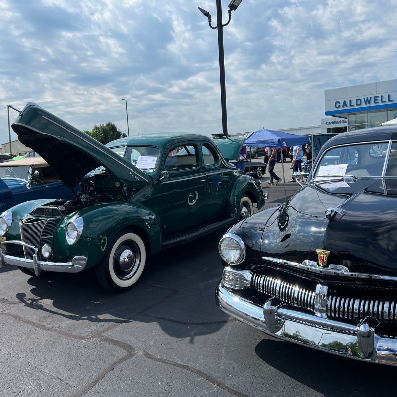 Two vintage cars at a car show, one dark green with hood open and one black polished sedan.