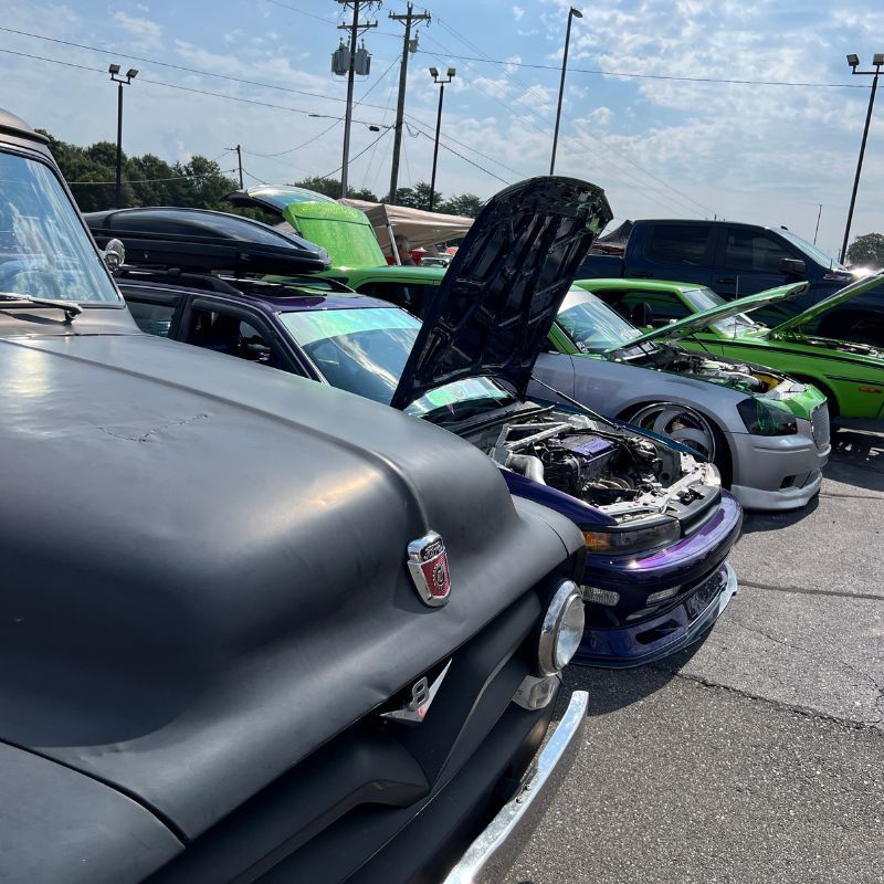 Classic cars parked at a show, including a matte black truck and a purple car with its hood open.