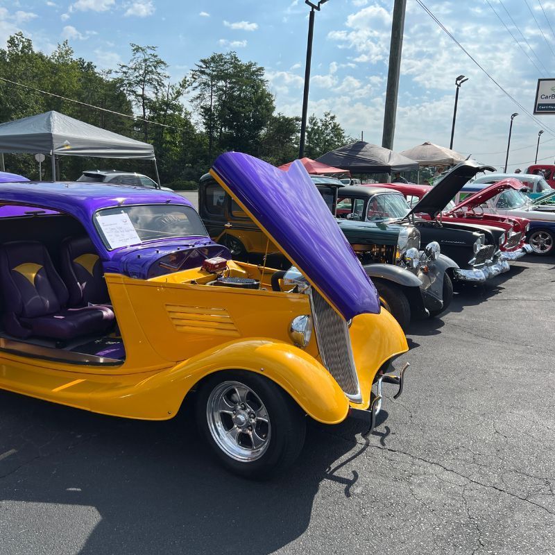 Purple and yellow classic custom car at an outdoor car show, hood open, with several cars in the background