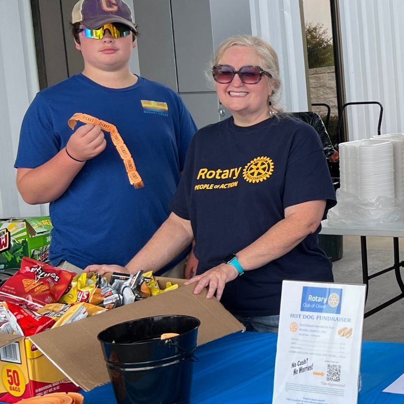 Two people in blue shirts standing behind a table of snacks and a sign at a Rotary event.