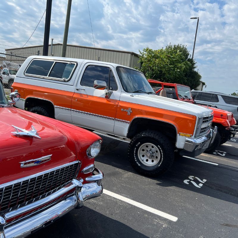 Classic cars parked in a lot, including a silver-and-orange SUV and a red vintage car in the foreground