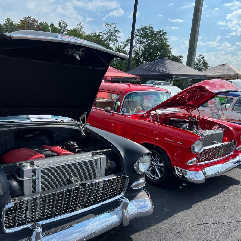 Classic cars with open hoods at an outdoor car show, including a black and red vintage Chevrolet.