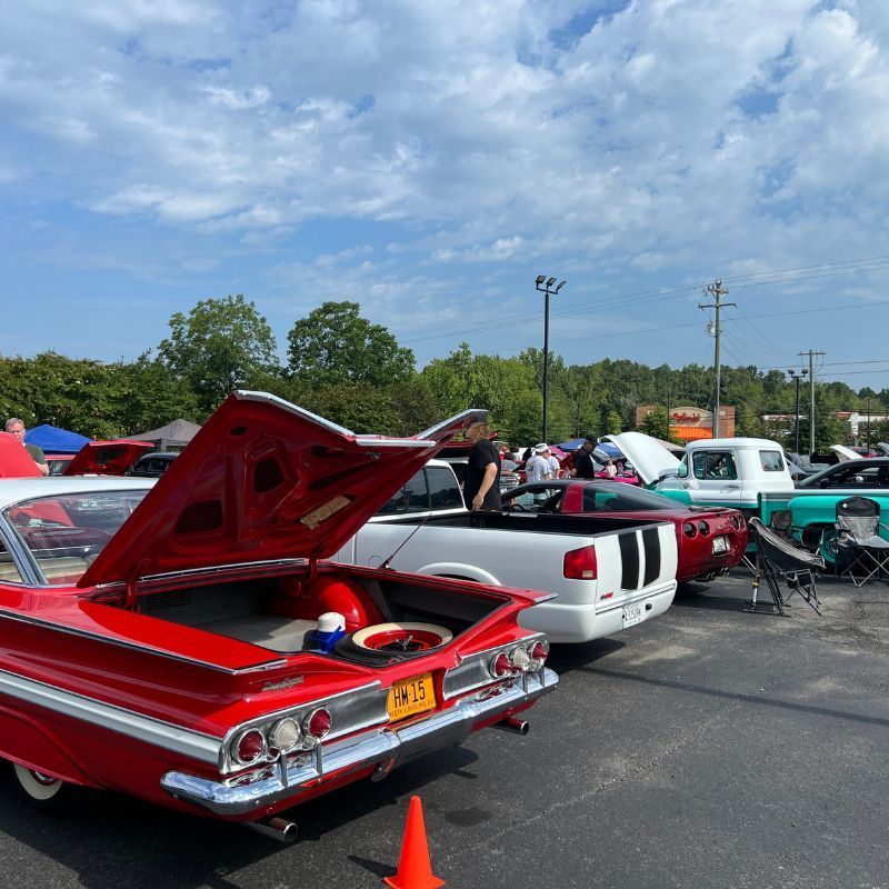 Classic car show with a red convertible, trunk open, and other vintage cars parked under a blue sky