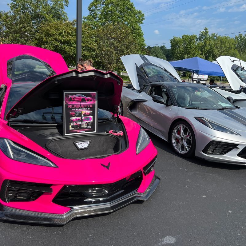 Bright pink and silver sports cars with open hoods at a car show, one with a display screen in front.