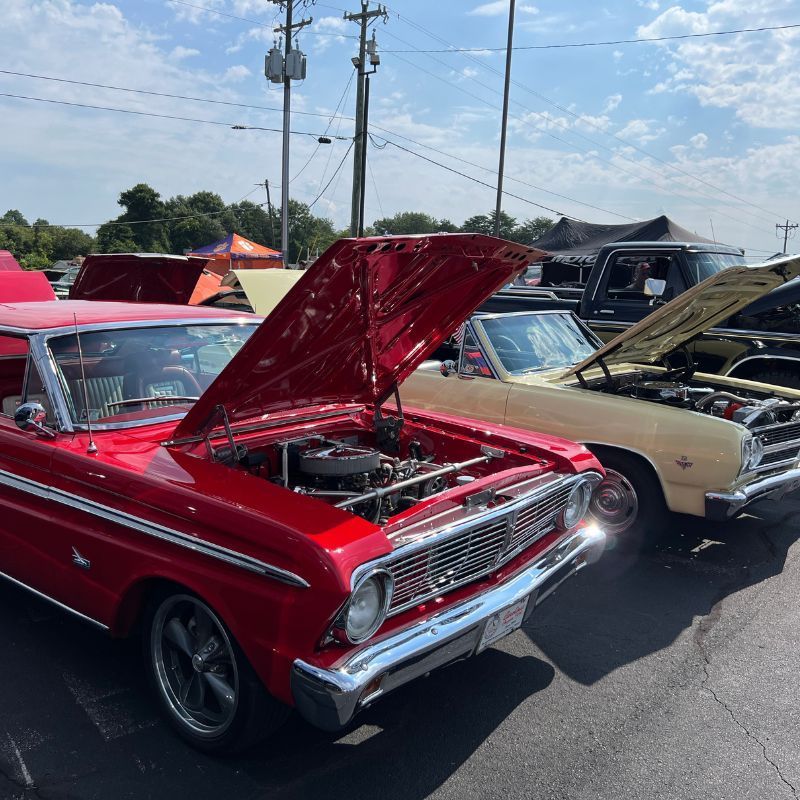 Classic red car with hood open at an outdoor car show, parked beside beige and black vintage cars.