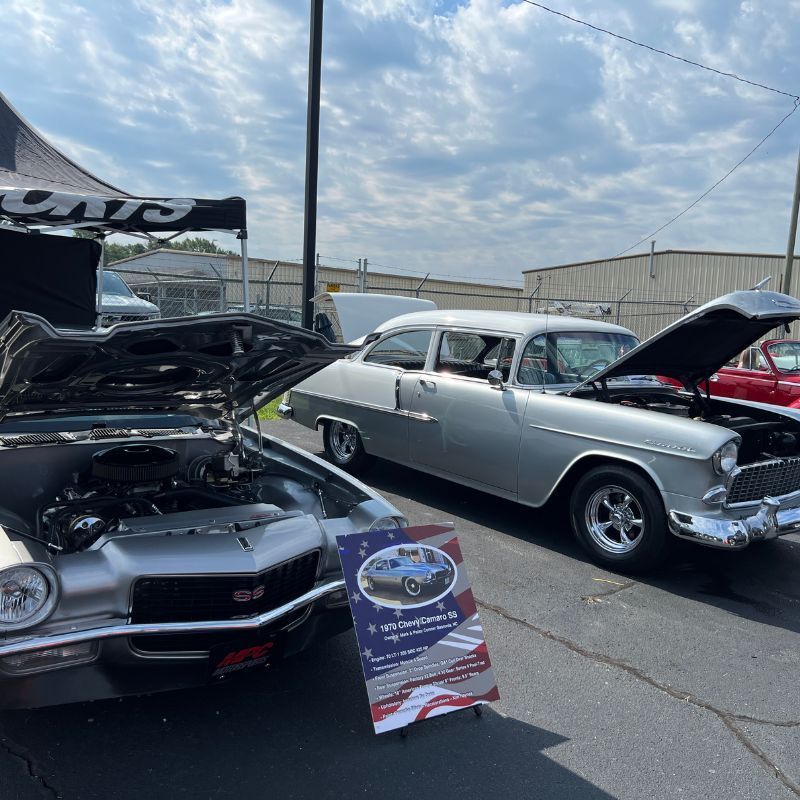 Classic cars on display at an outdoor show, including a silver 1950s coupe with its hood open.
