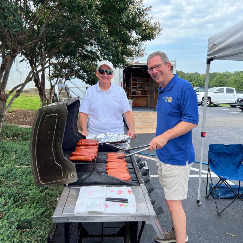 Two men grilling hot dogs outdoors beside a parking lot, smiling near a barbecue smoker.