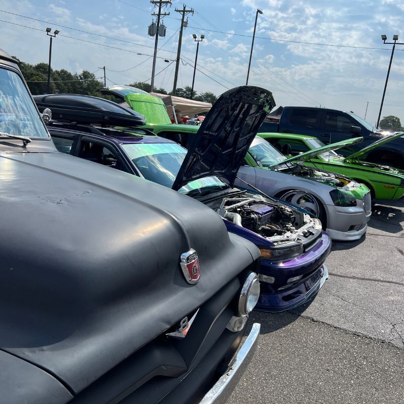 Classic cars with hoods open at an outdoor car show, including a matte black car in the foreground
