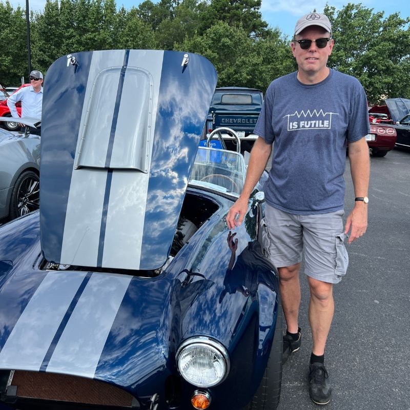 Man standing beside a blue classic race car with its hood open at an outdoor car show