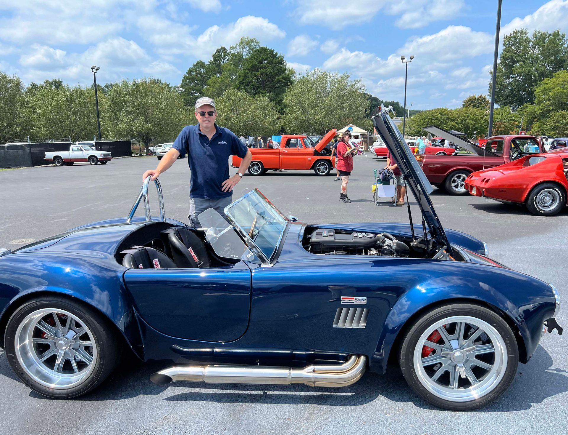 Blue sports car with a man standing beside it in a parking lot, with other cars in the background