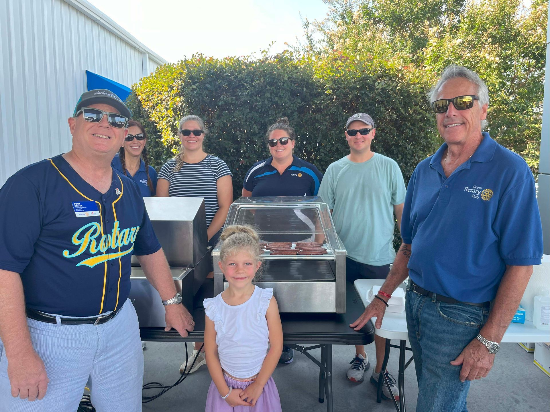 Six people posing around a grill outdoors, with a child seated in front, wearing casual summer clothes.