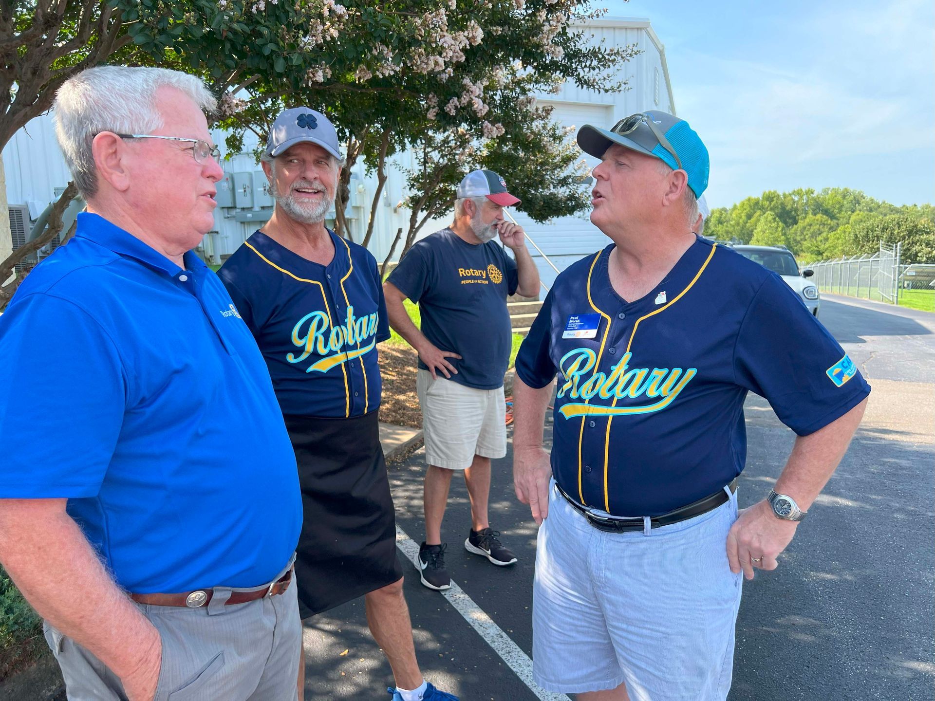 Four men chatting outdoors on a road, wearing blue and navy shirts and caps on a sunny day