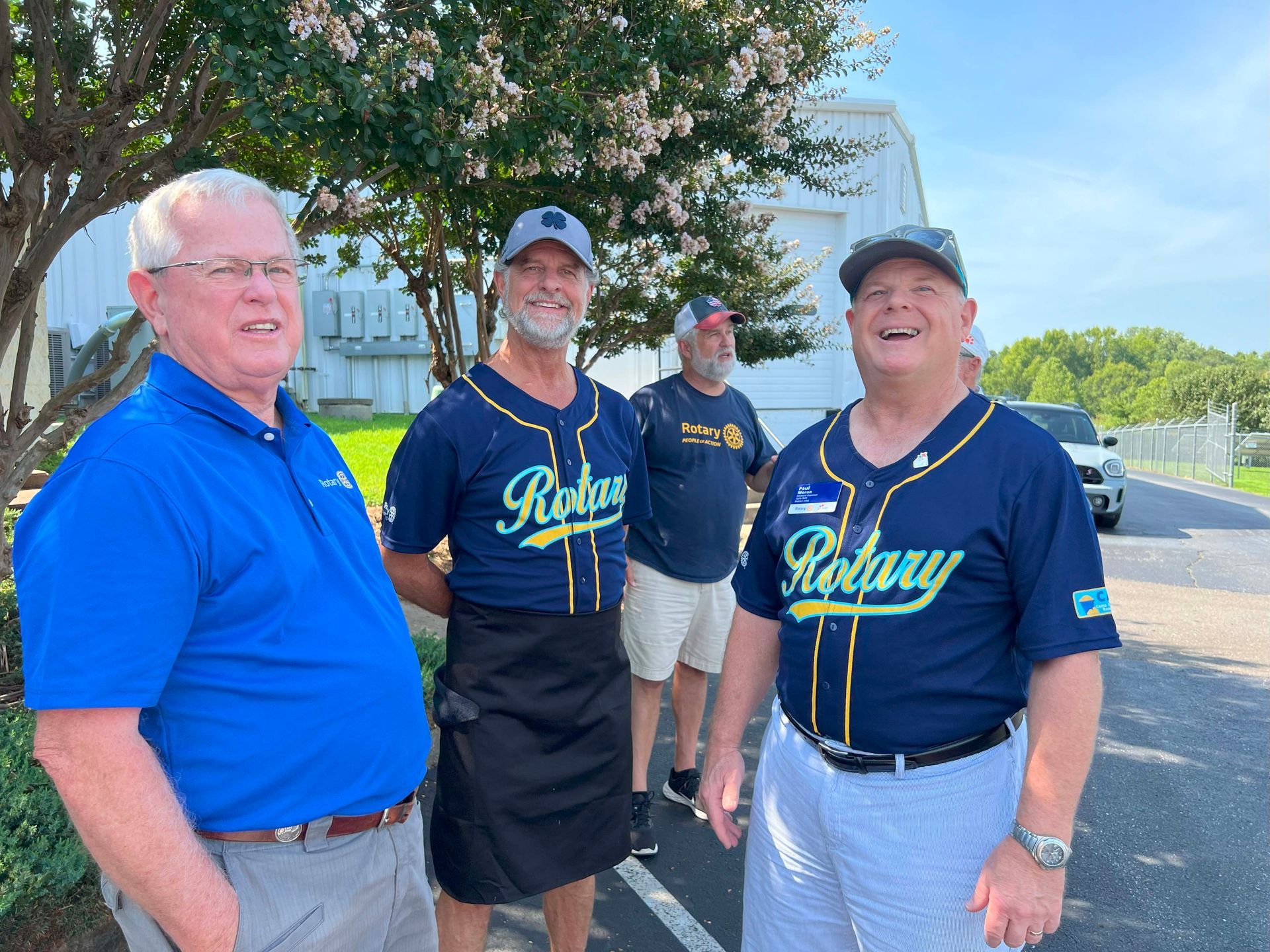 Three smiling men in blue Rotary shirts standing outdoors near a road and trees