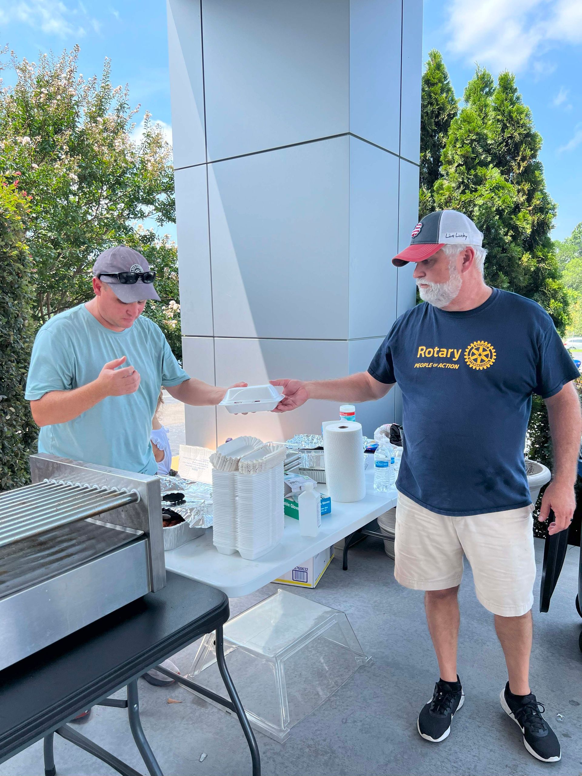 Two men exchanging a cup at an outdoor food table beside a grill and beverage station.