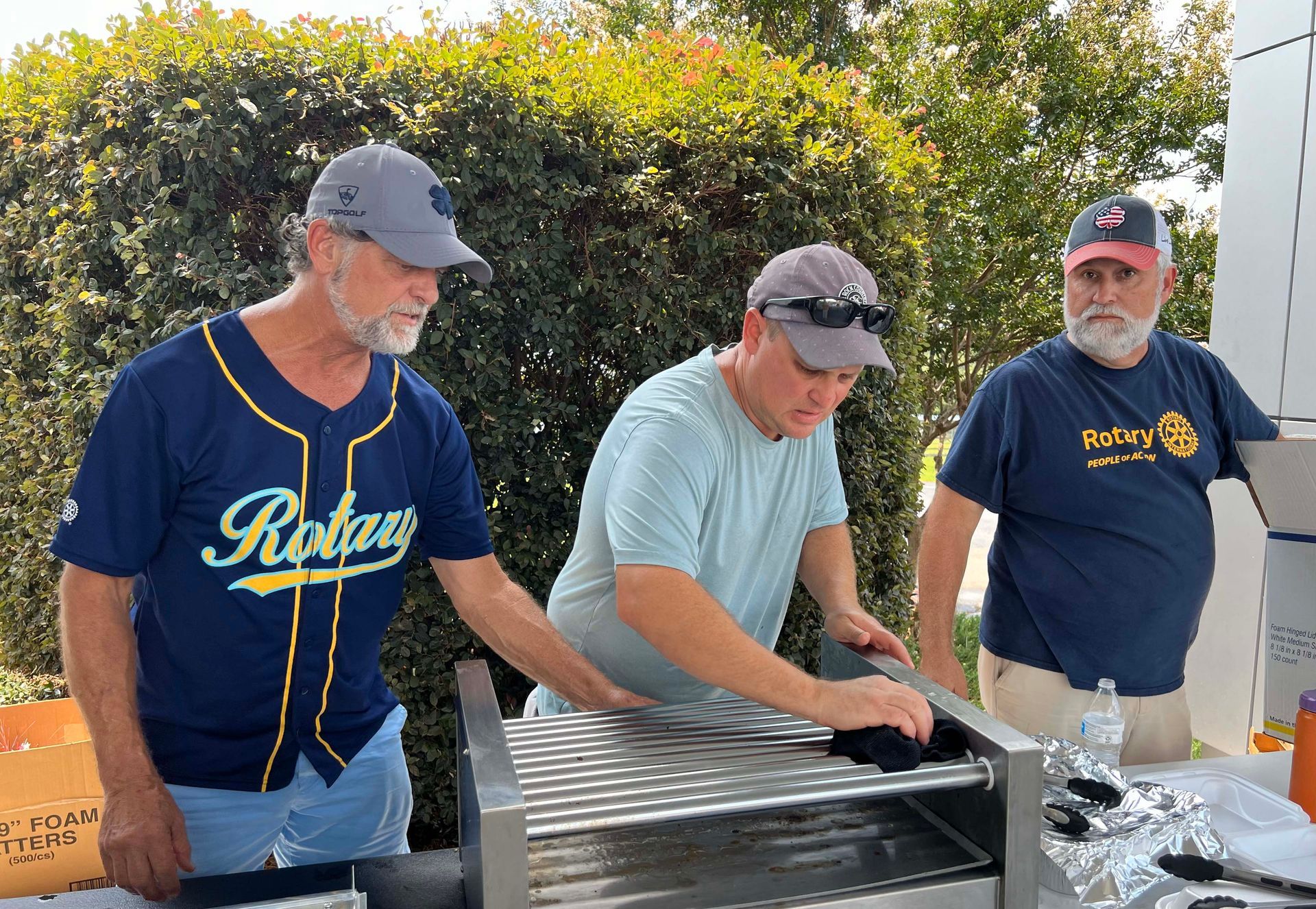 Three men in Rays shirts prepare food at an outdoor grill table.