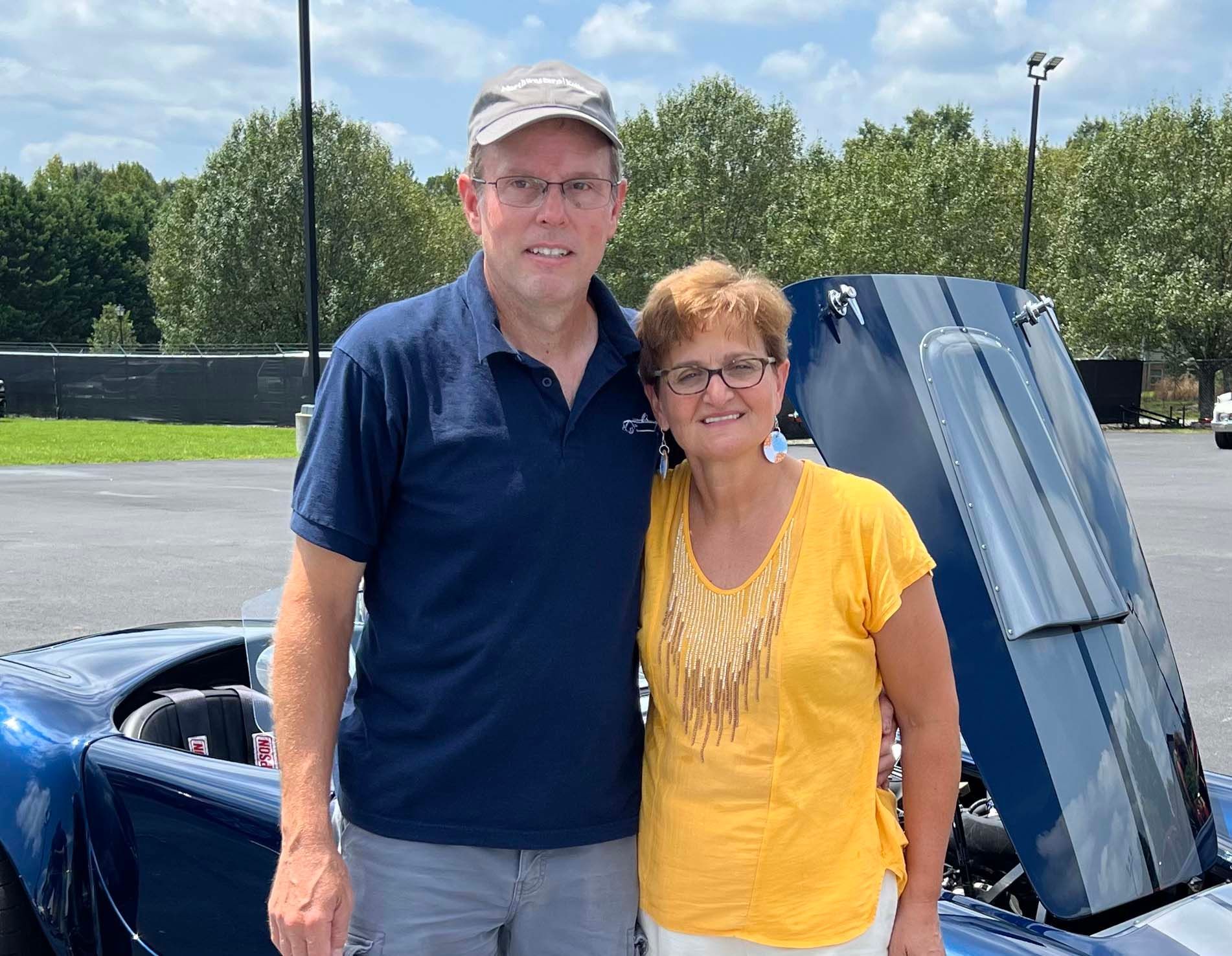 Two people posing beside a blue sports car outdoors, with the hood raised.