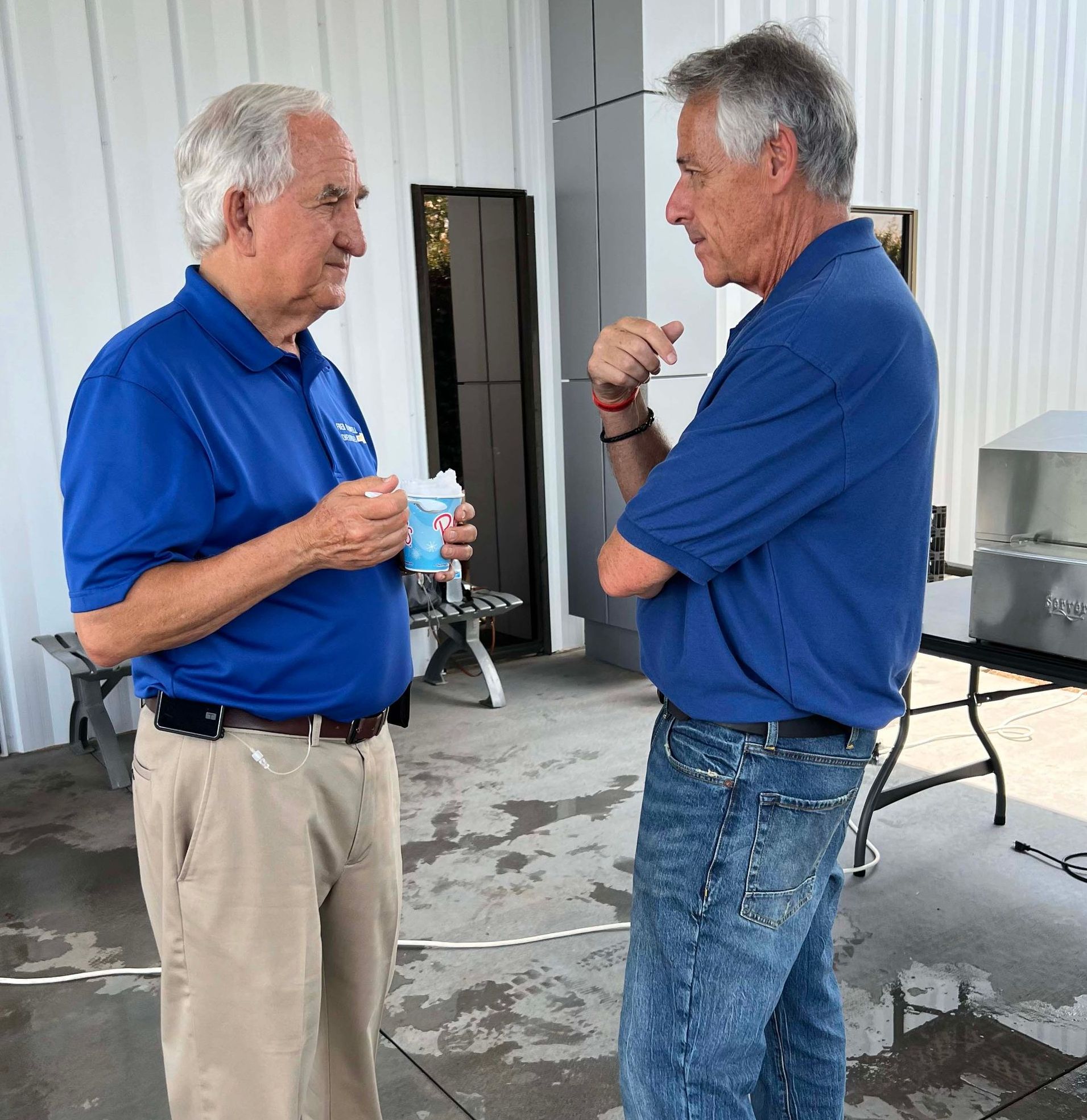 Two men in blue polos talk in a bright patio or garage area, one holding a cup and the other gesturing.