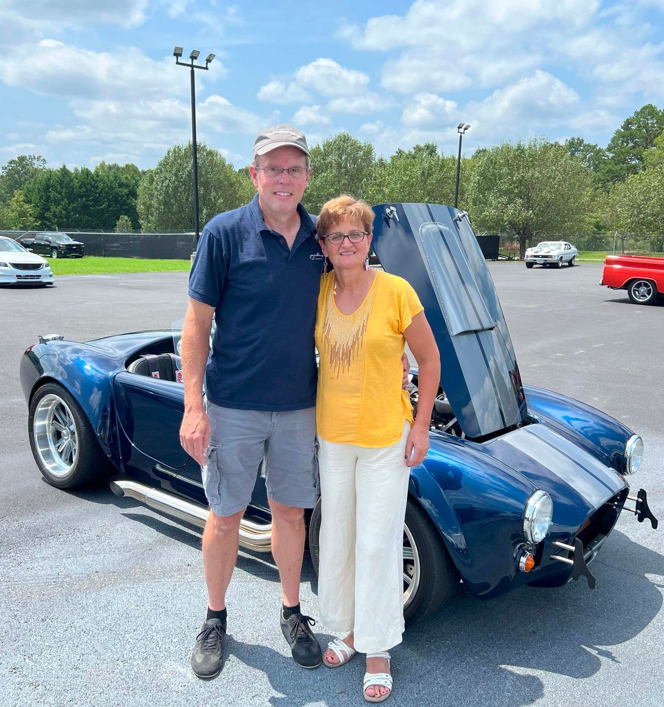 Two people standing beside a blue sports car with its hood open in a parking lot.