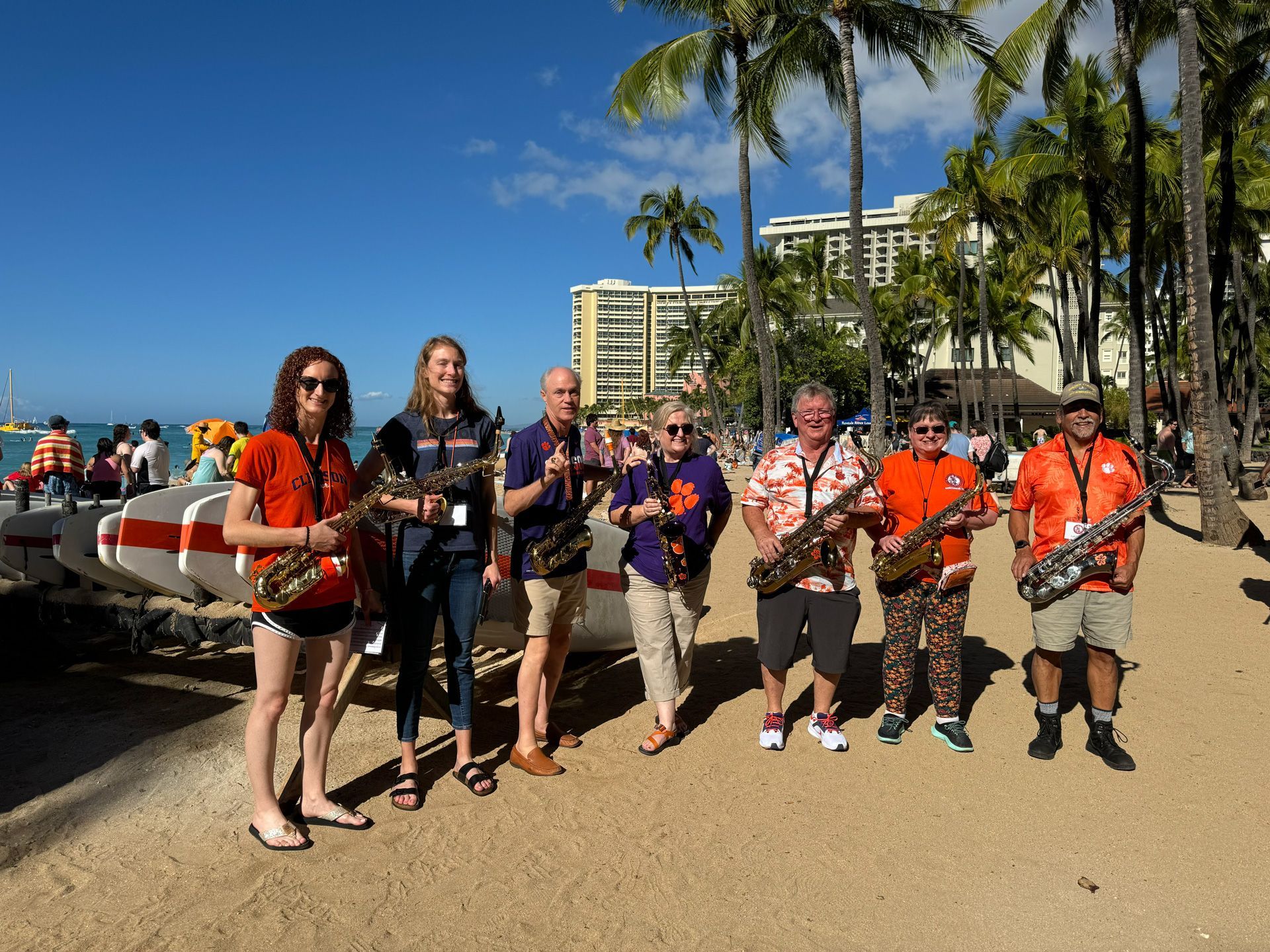 Group posing with ukuleles on a sunny beach, palm trees and resort buildings in the background.