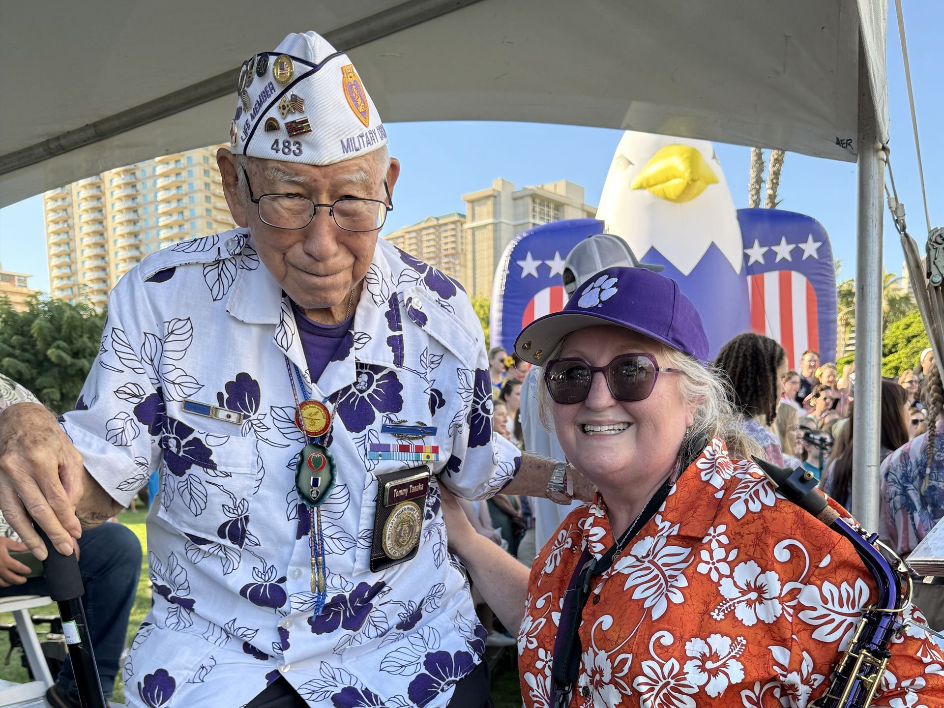 Two smiling people in floral shirts and veteran caps posing under a canopy at an outdoor event