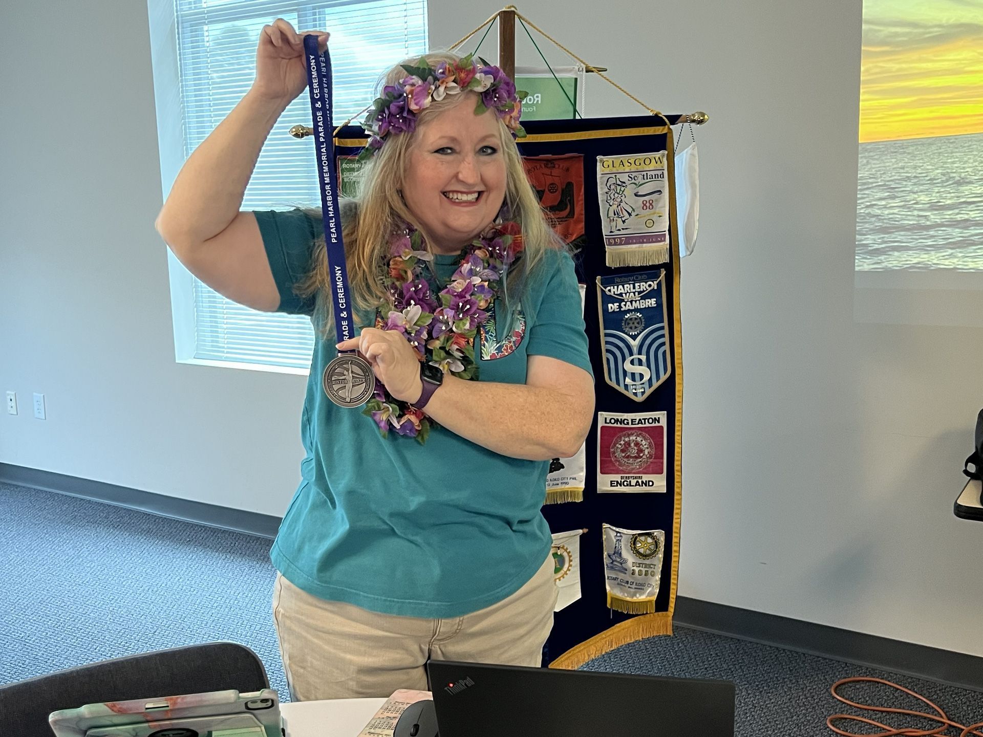 Smiling person holding a ribboned award banner indoors, wearing teal shirt and floral lei.