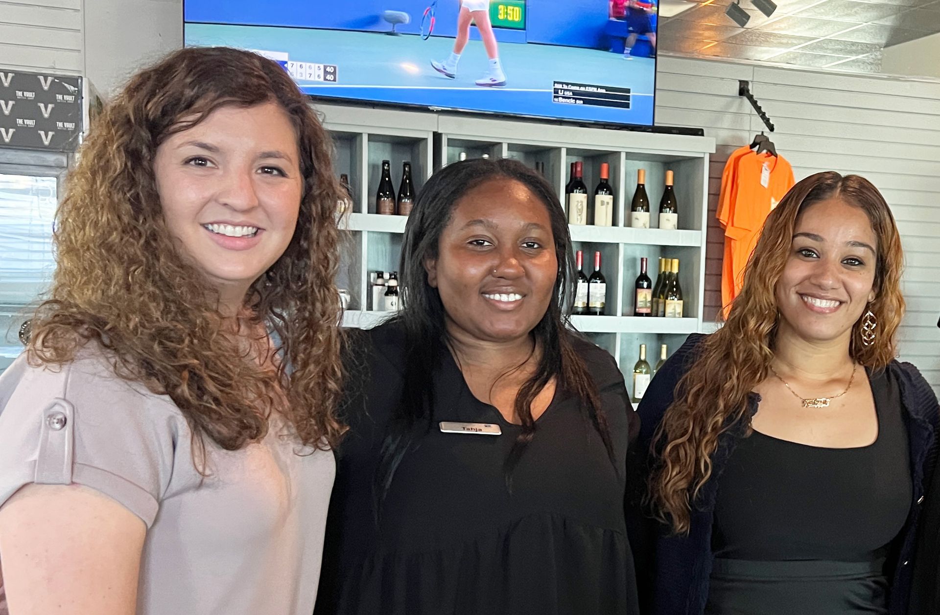 Three women smiling in front of a bar with bottles and TV screens behind them