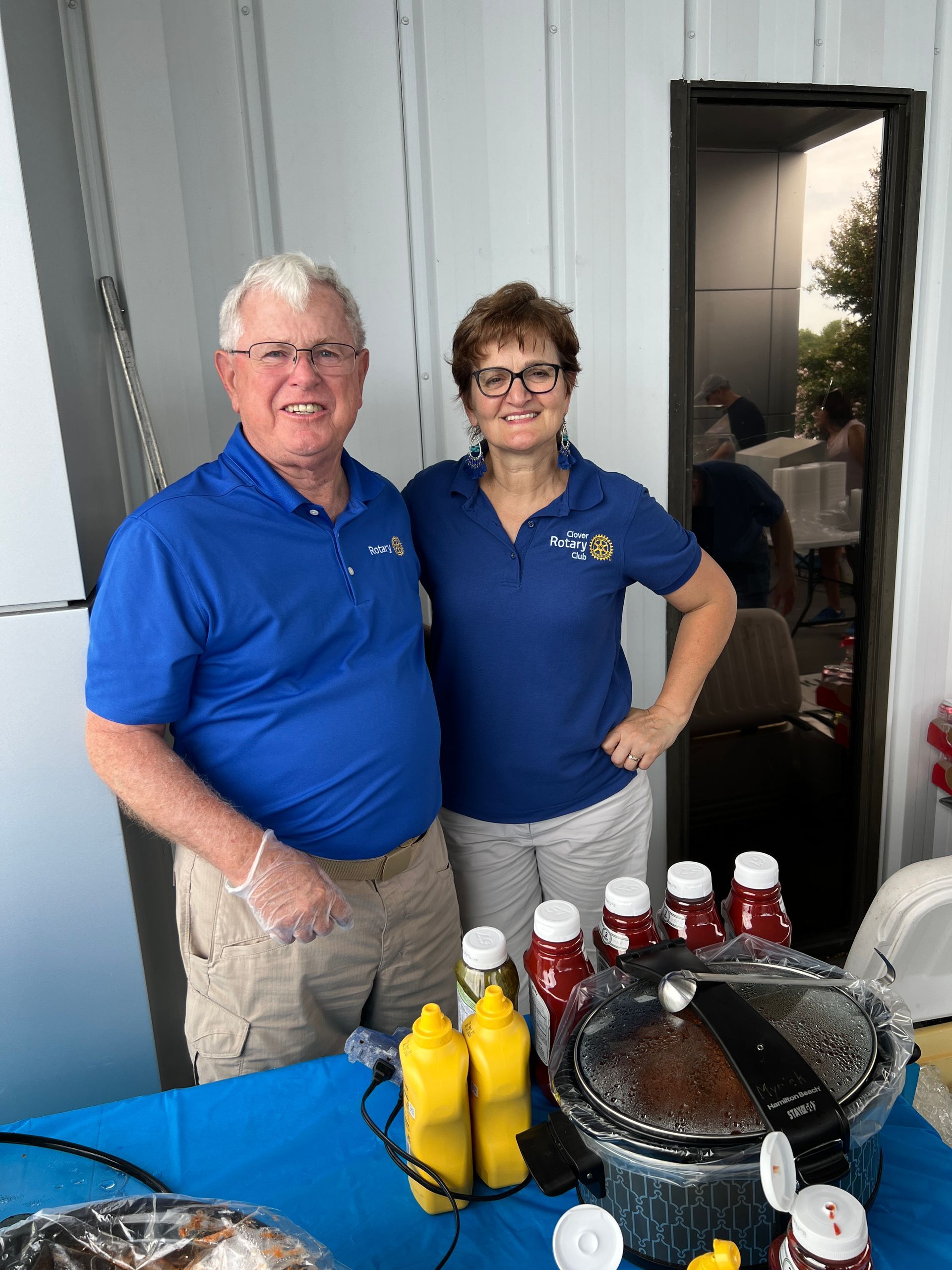 Two people in blue shirts stand behind a table with drinks and a covered grill.