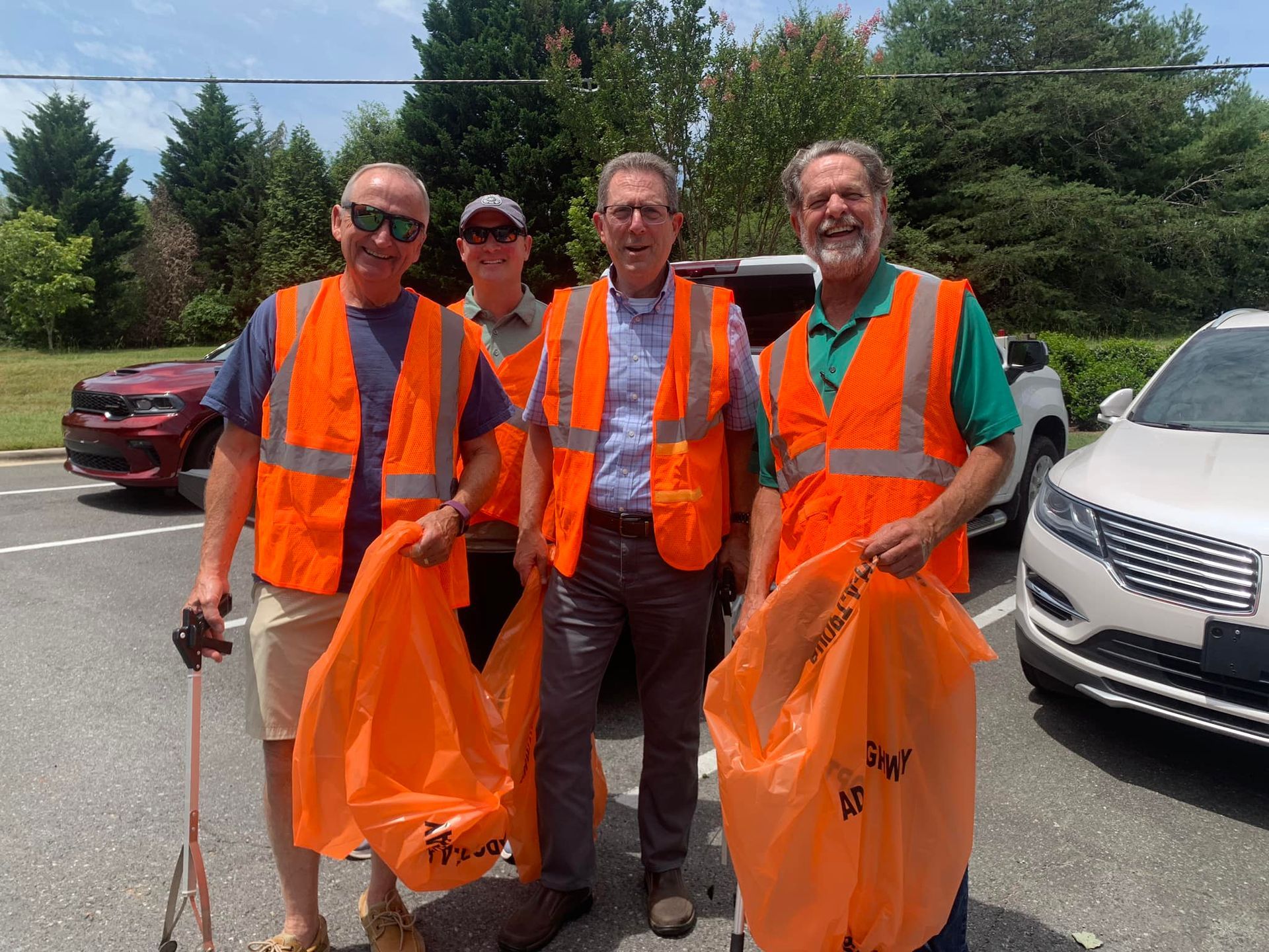 Four road workers in orange safety vests holding orange trash bags on a roadside.