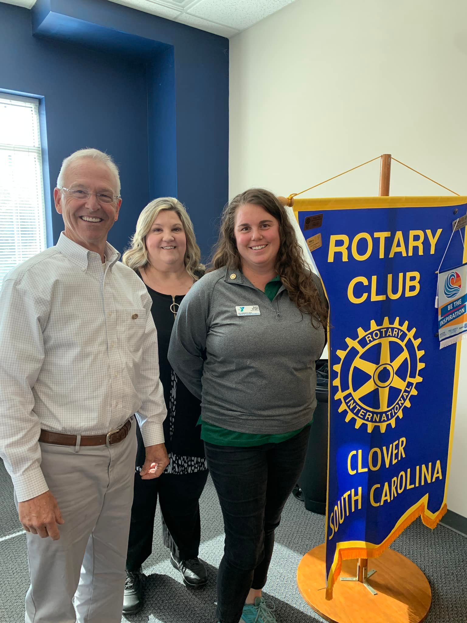 Three people stand beside a blue-and-gold Rotary Club banner in a bright room.