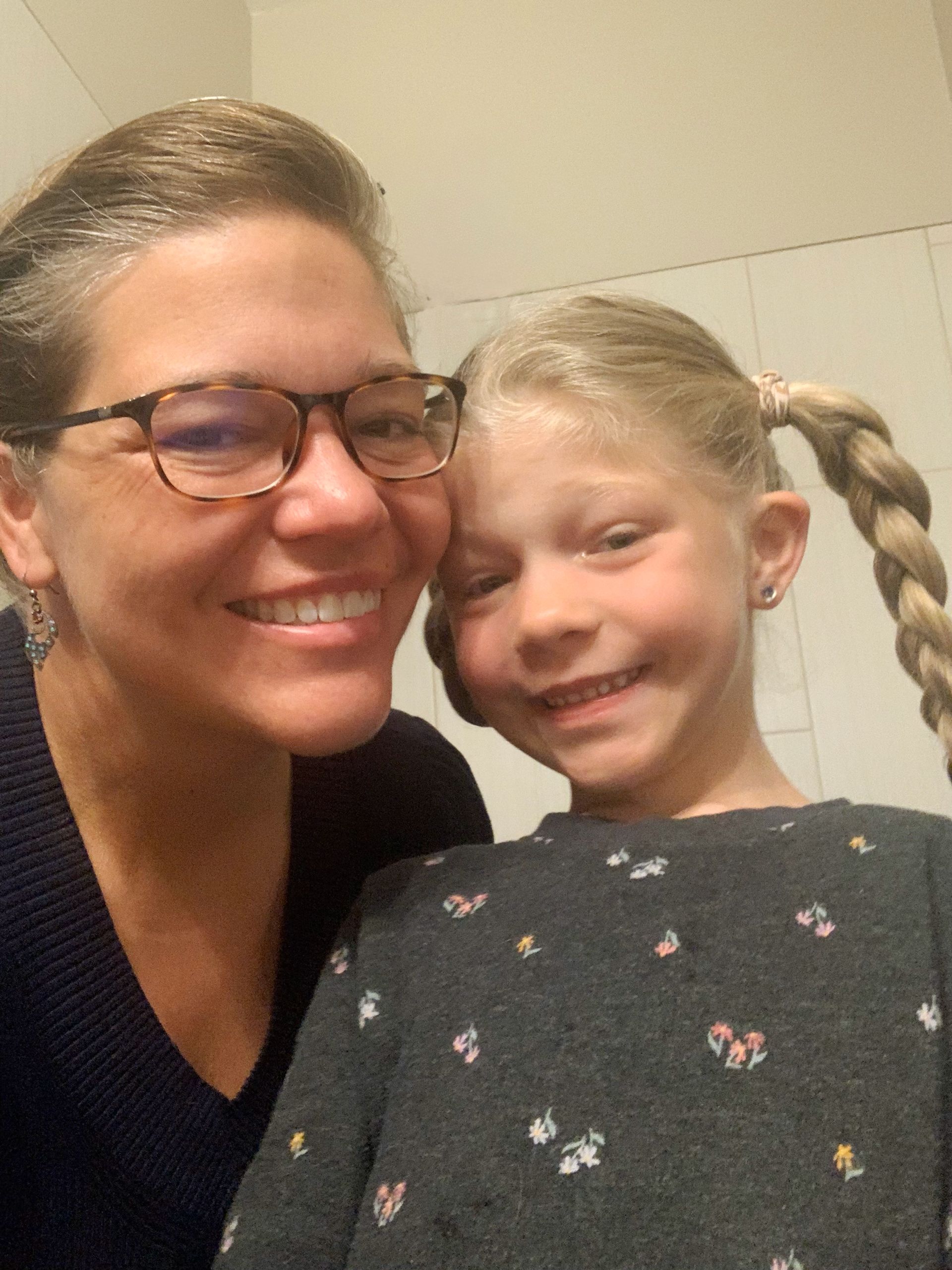 Two smiling people posing close together indoors, one wearing glasses and the other with braided hair.