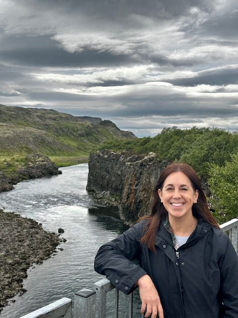Woman smiling by a river gorge under cloudy skies, with rocky cliffs and green hills in the background