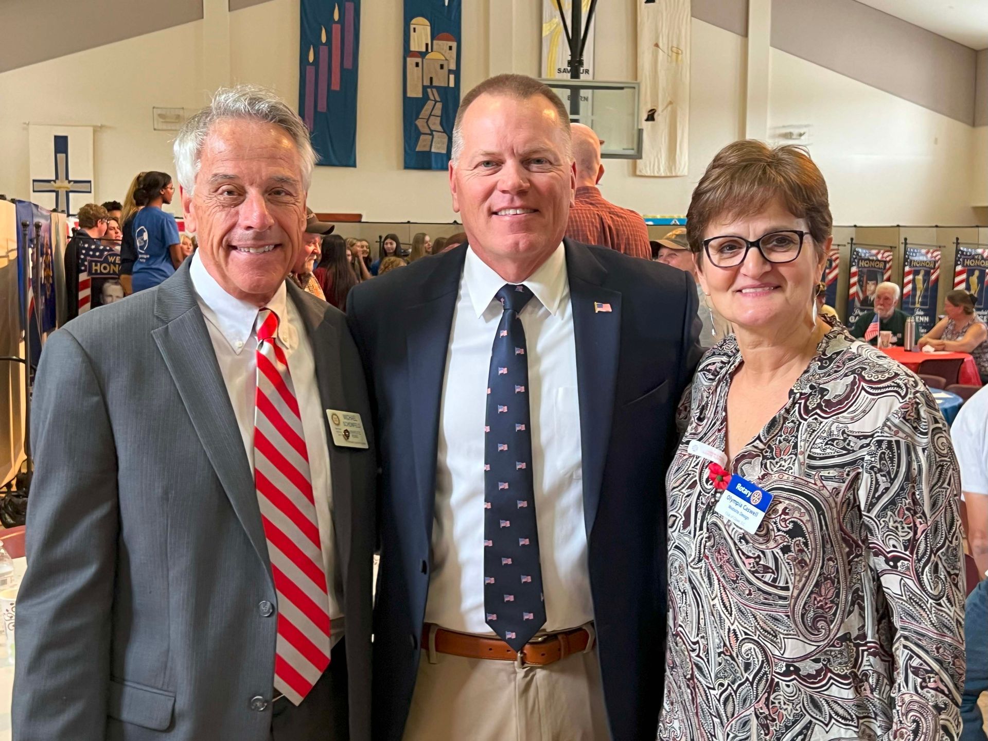 Three people posing indoors at a formal event, with banners and a crowd in the background.