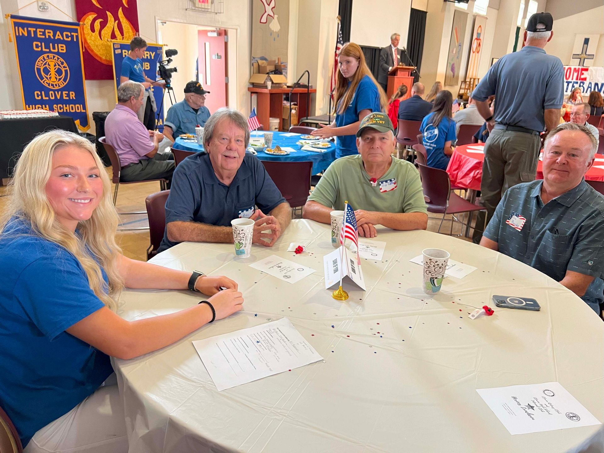 People seated at a round table in a busy community hall, with papers, drinks, and a card game in progress.