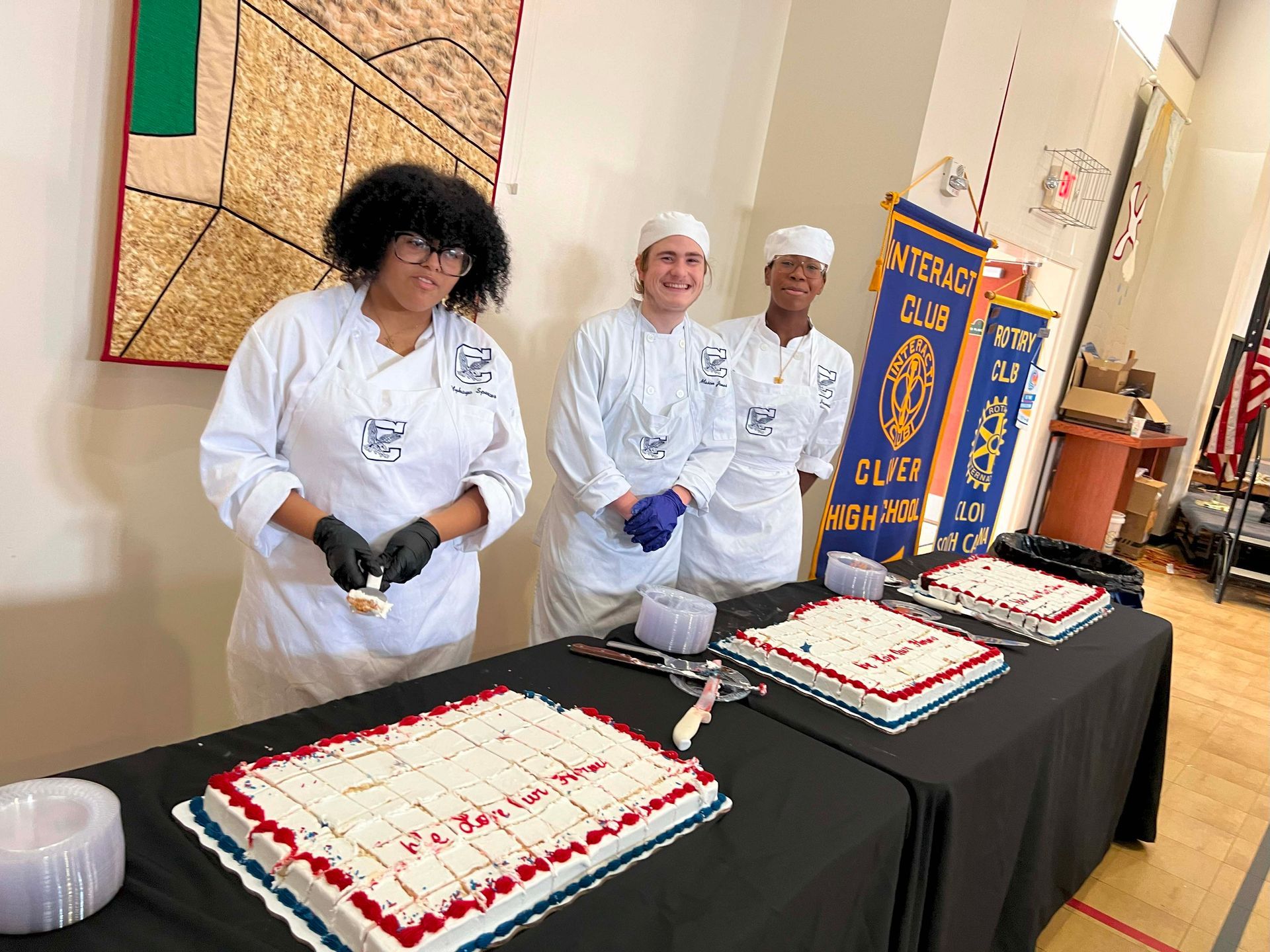 People in white uniforms stand behind decorated cakes at a table in a hallway.