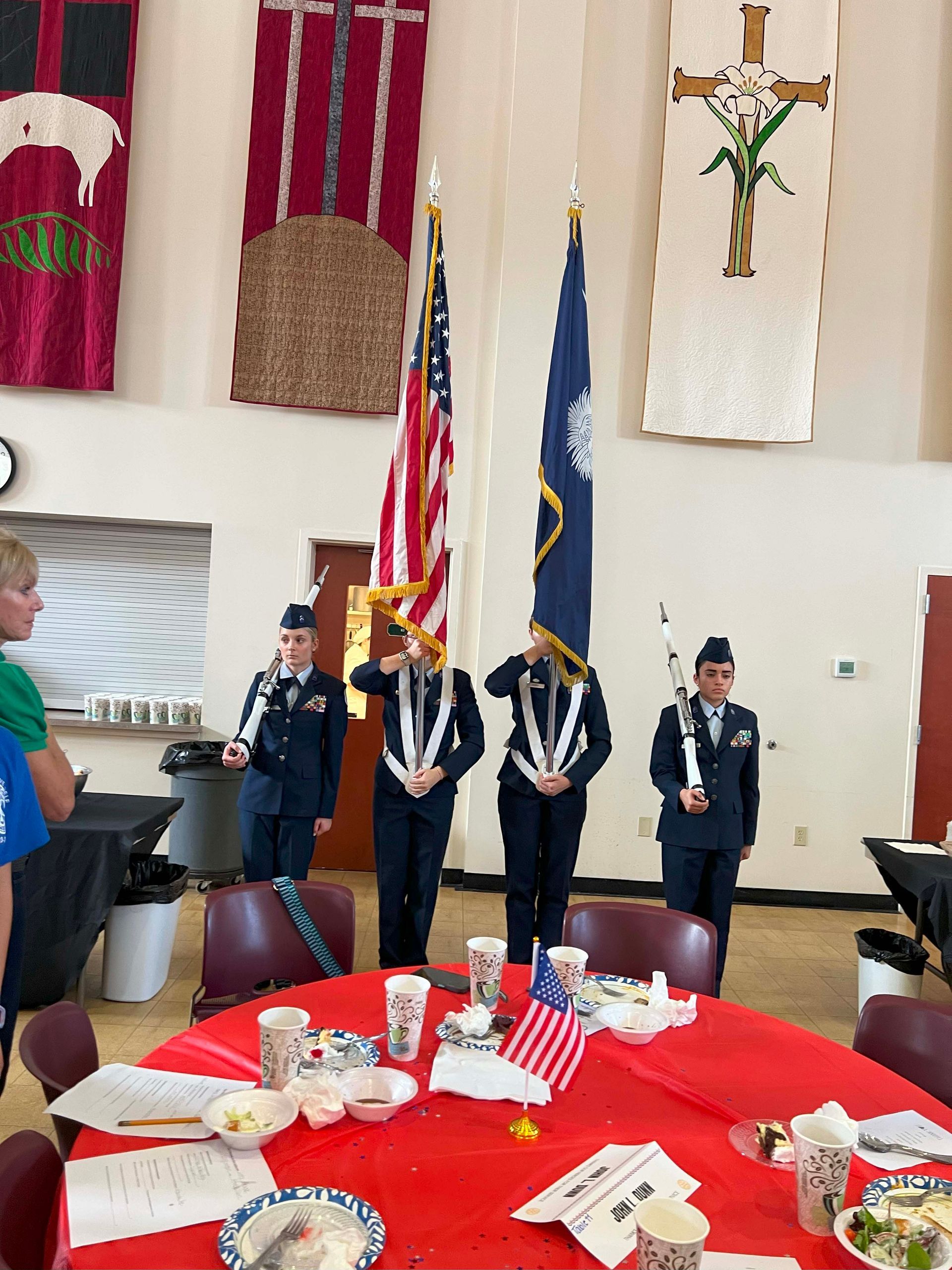 Color guard holding flags in a church hall beside a red table with small American flags and refreshments