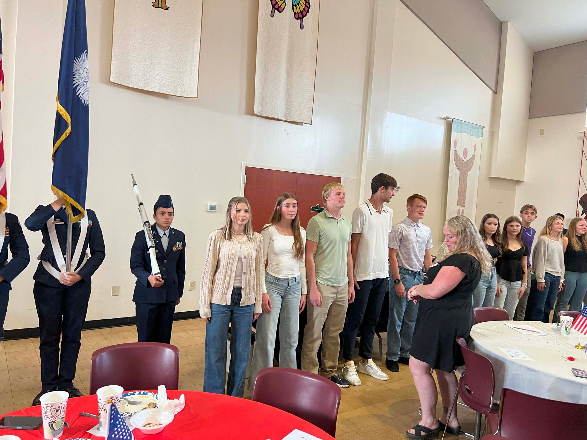 People standing in a church or hall beside tables set for a gathering, with flags and wall banners in the background.