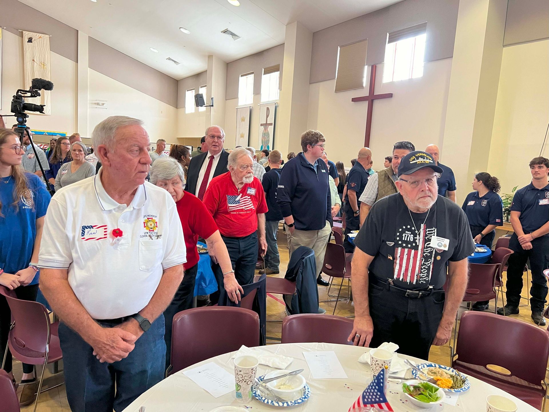People standing in a church hall during a gathering, with tables set in the foreground and a cross on the wall.