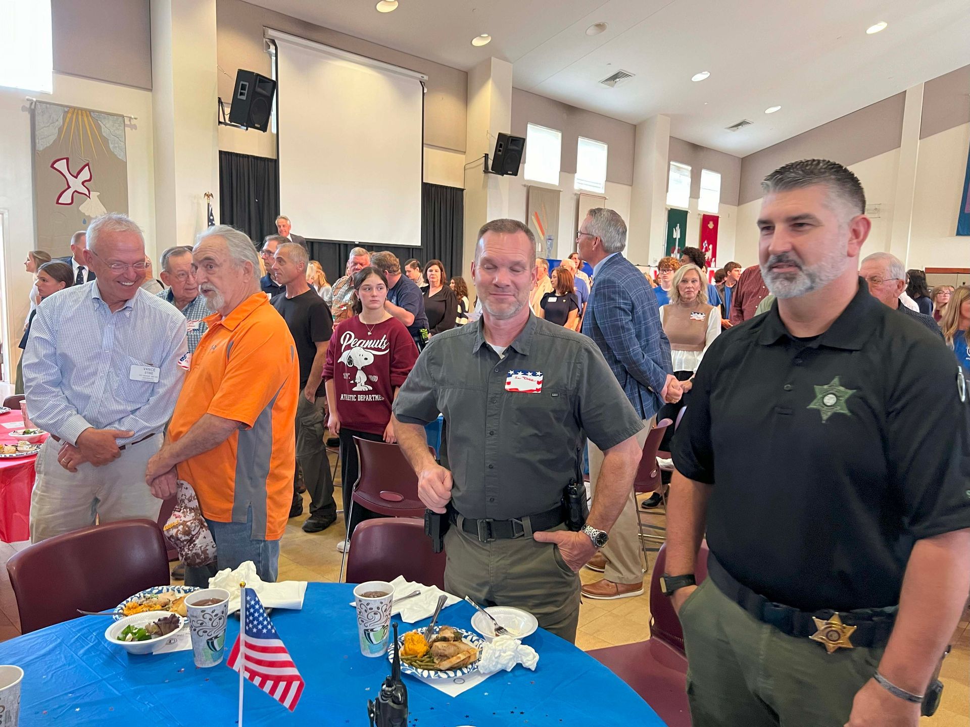 People standing around a table with food and small American flags in a bright indoor hall.