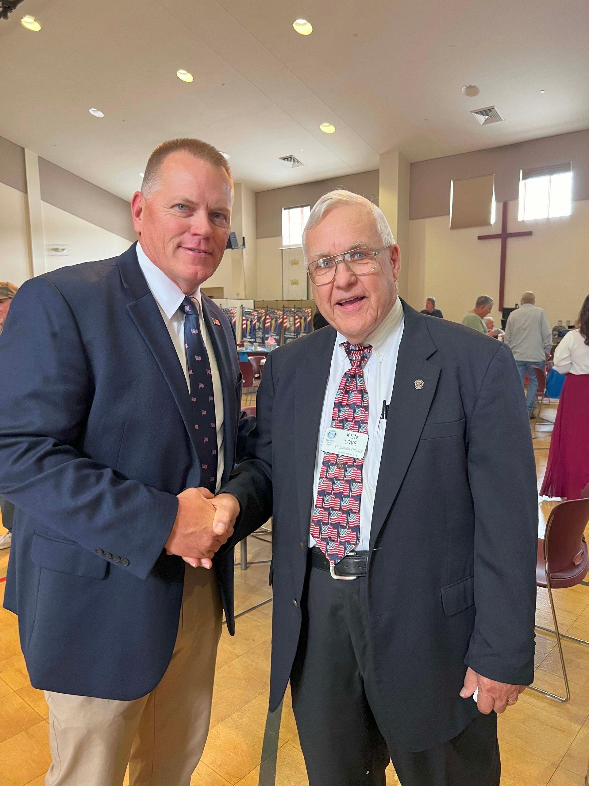 Two men in suits shaking hands in a church hall with chairs and people in the background.