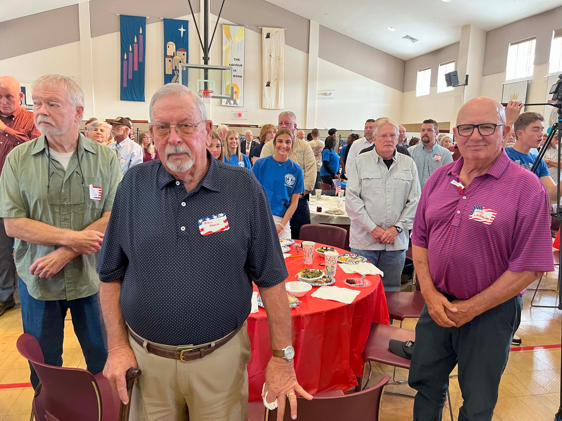 People gathered in a hall around red tablecloths, standing and facing the camera.