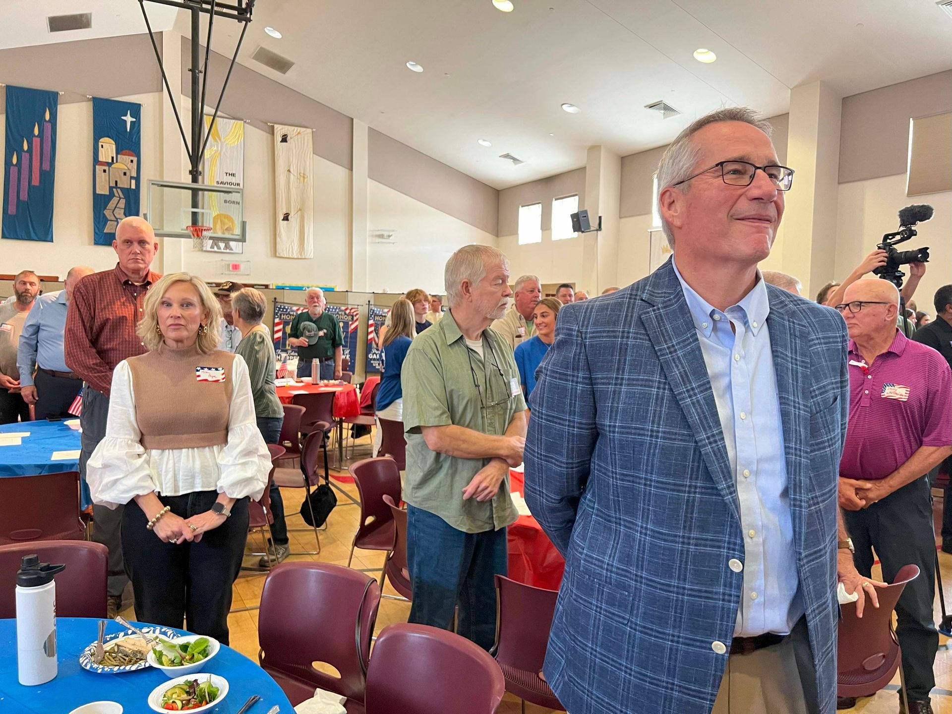People standing in a bright community hall, with a smiling man in a blue plaid jacket in the foreground.