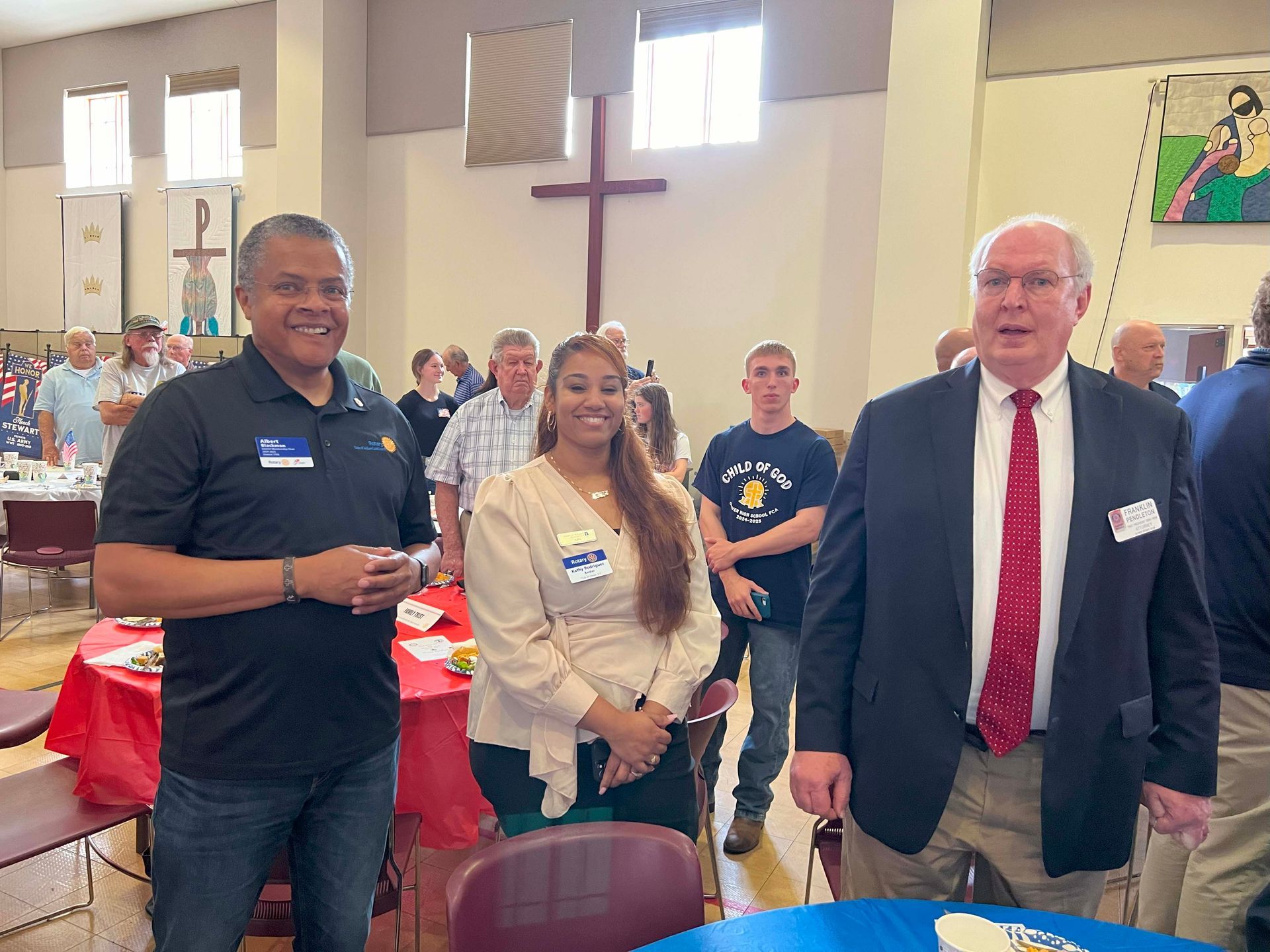 Three people standing in a church hall, smiling near a large cross and seated attendees.