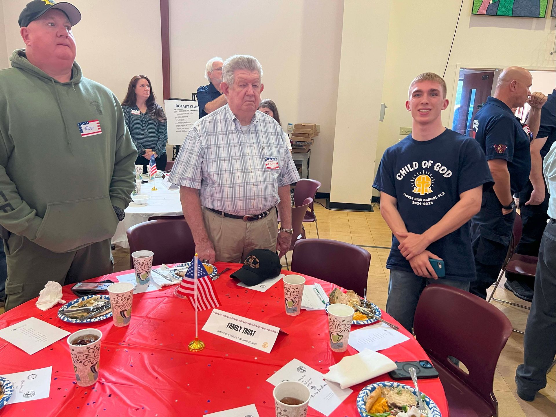 People standing around a red table at an indoor event, with small flags and papers scattered on it