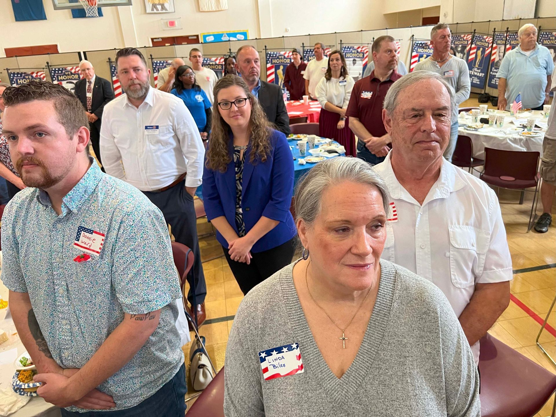 People standing in a bright meeting room with tables, facing forward, wearing name tags.