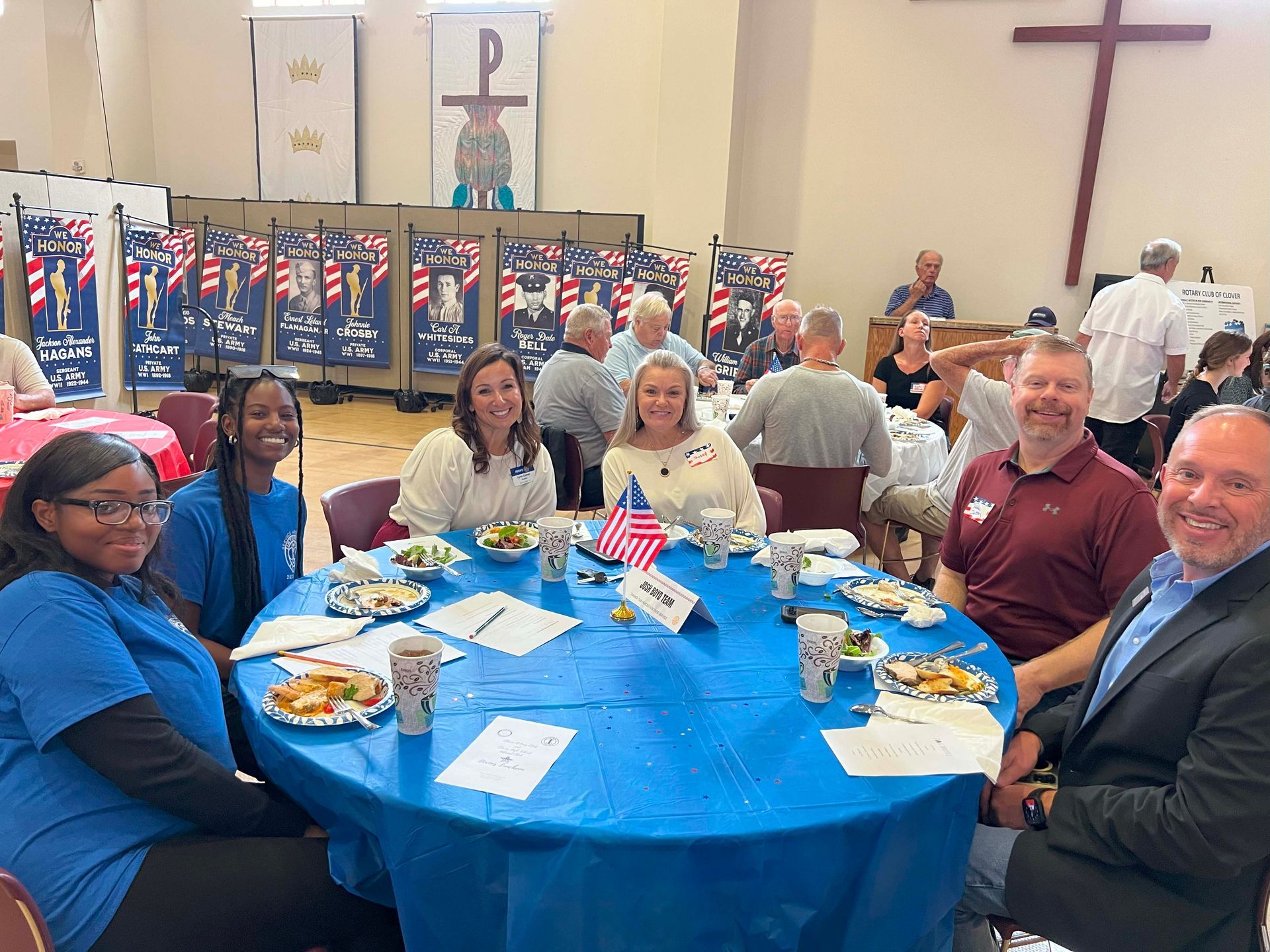 Group seated at a blue-covered banquet table in a hall with flags and a large cross in the background