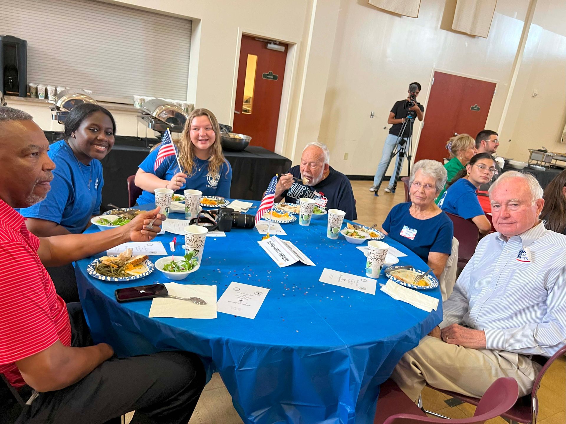 People seated around a blue-clothed banquet table, smiling and eating in a bright hall.