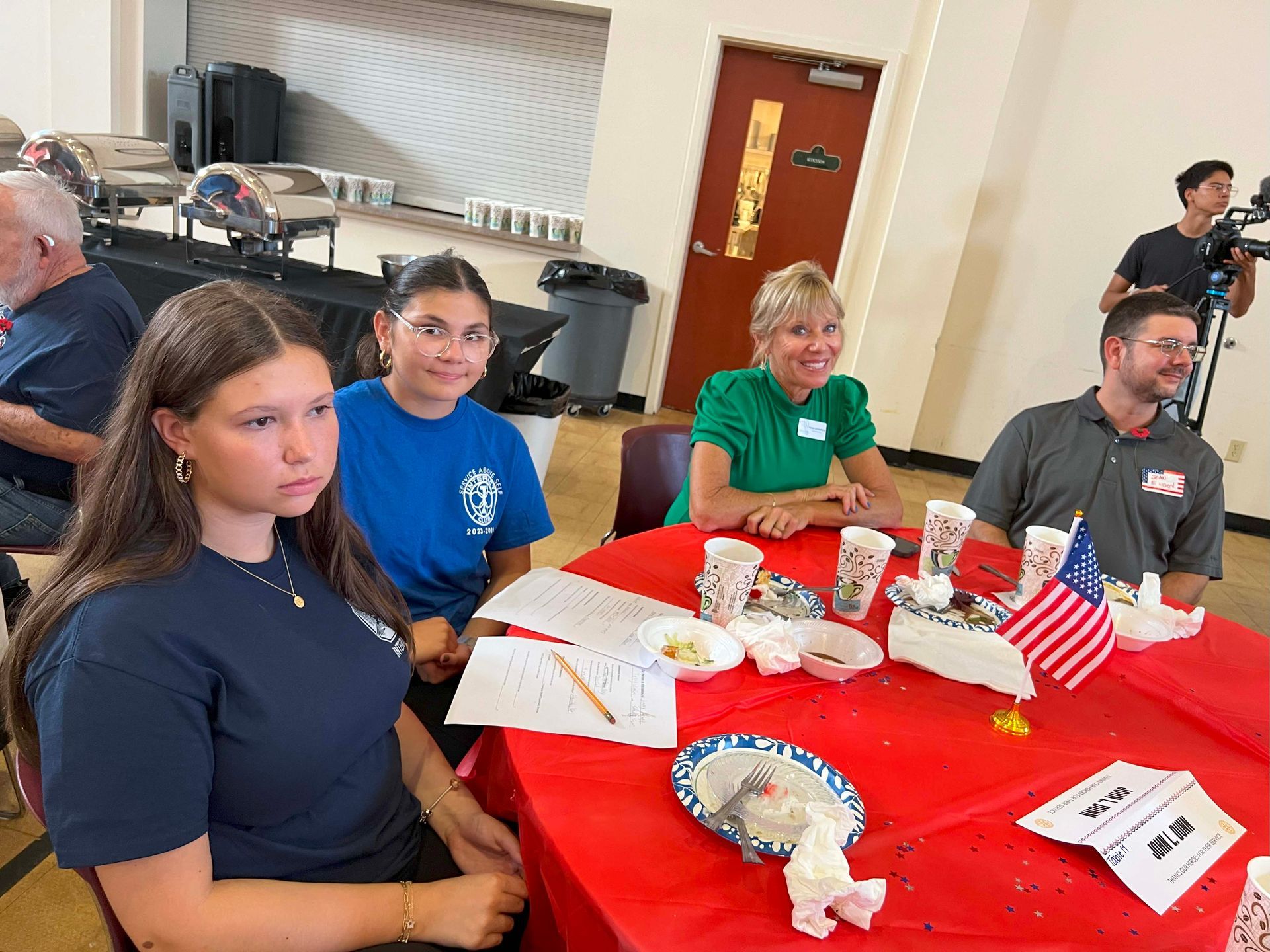 People seated at a red-covered table in a bright room, with papers, snacks, and patriotic decorations.