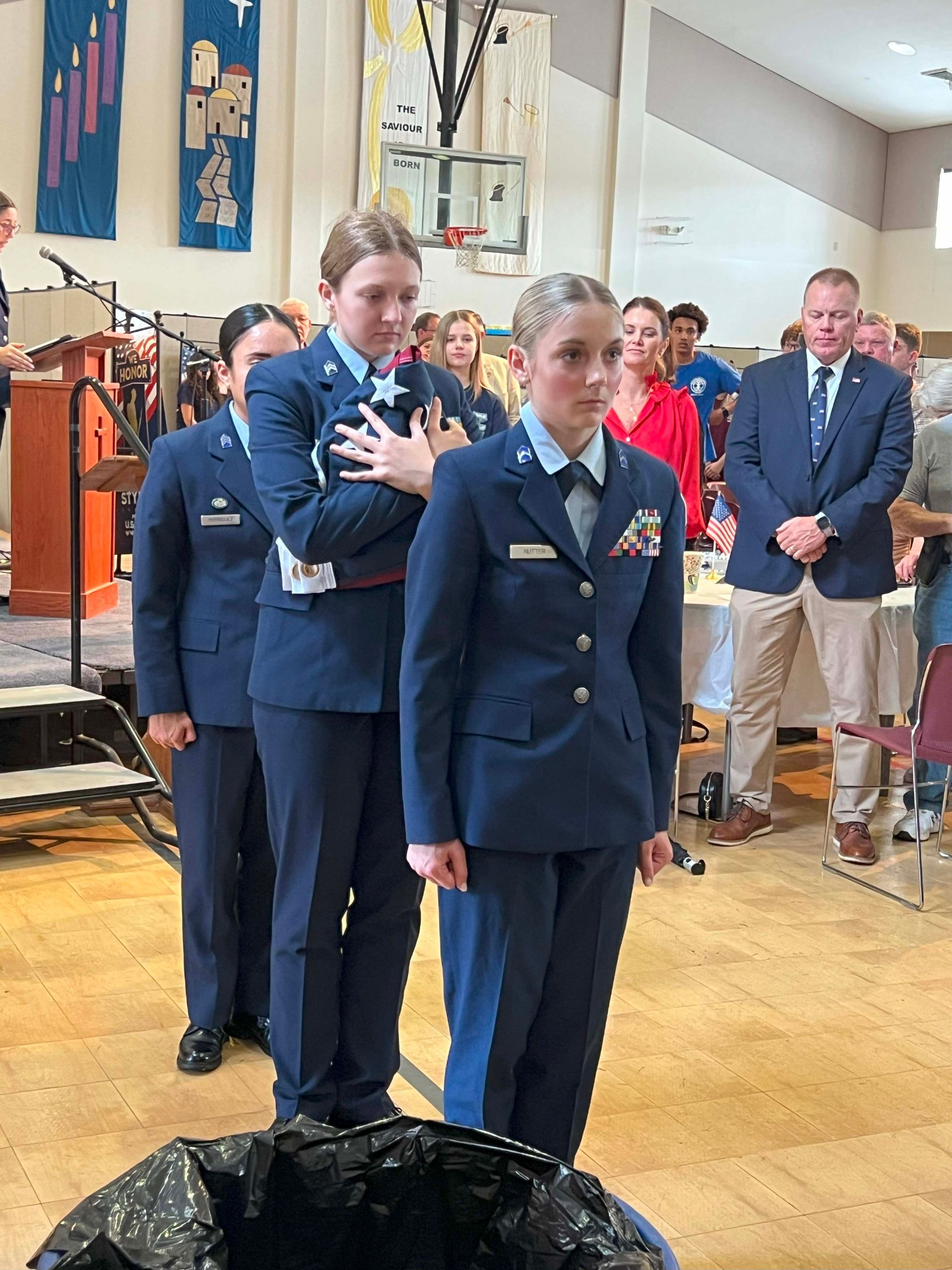 Two cadets in navy uniforms stand indoors at a formal ceremony, one with hand over heart.
