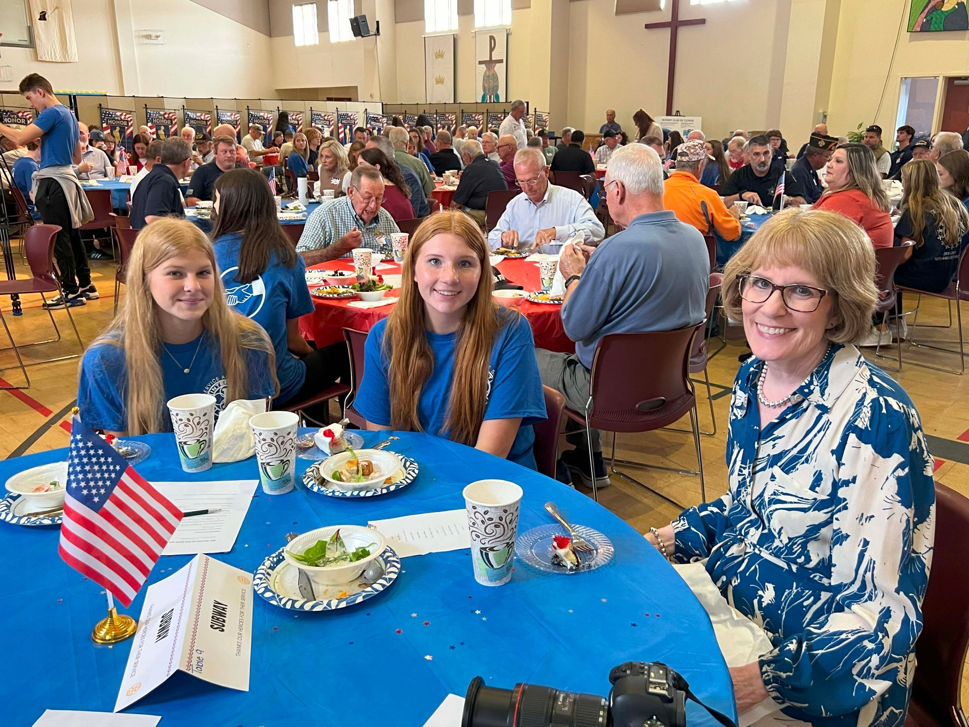 People seated at blue tables in a church hall during a community meal, with a cross on the wall.